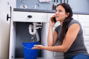 A woman on the phone as she indicates to a leaking pipe beneath a kitchen sink.